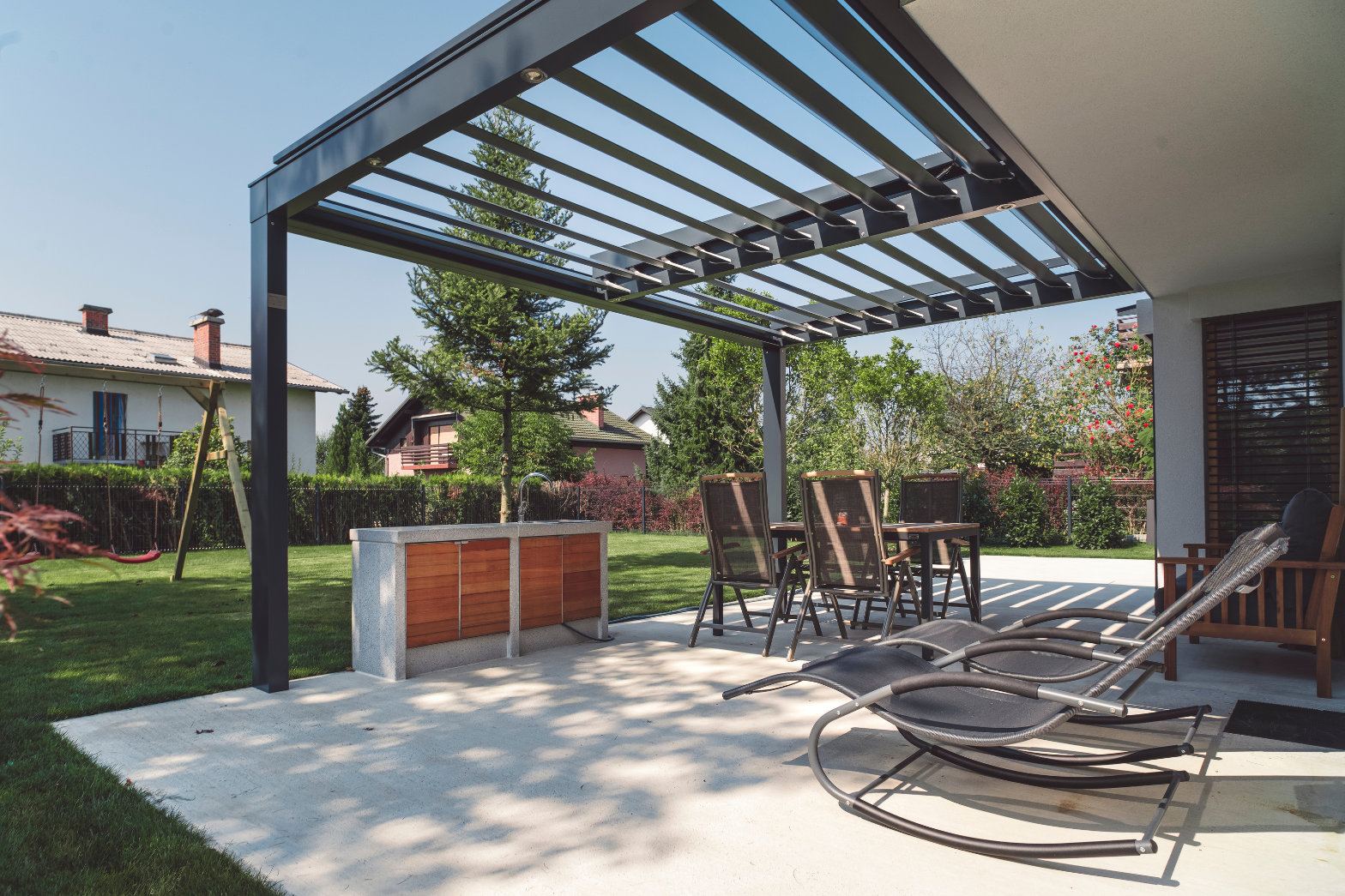 Moderne terrasse med pergola, utekjøkken og loungemøbler i hagen Moderne terrasse med pergola, utekjøkken, solstoler og spisegruppe i hage.